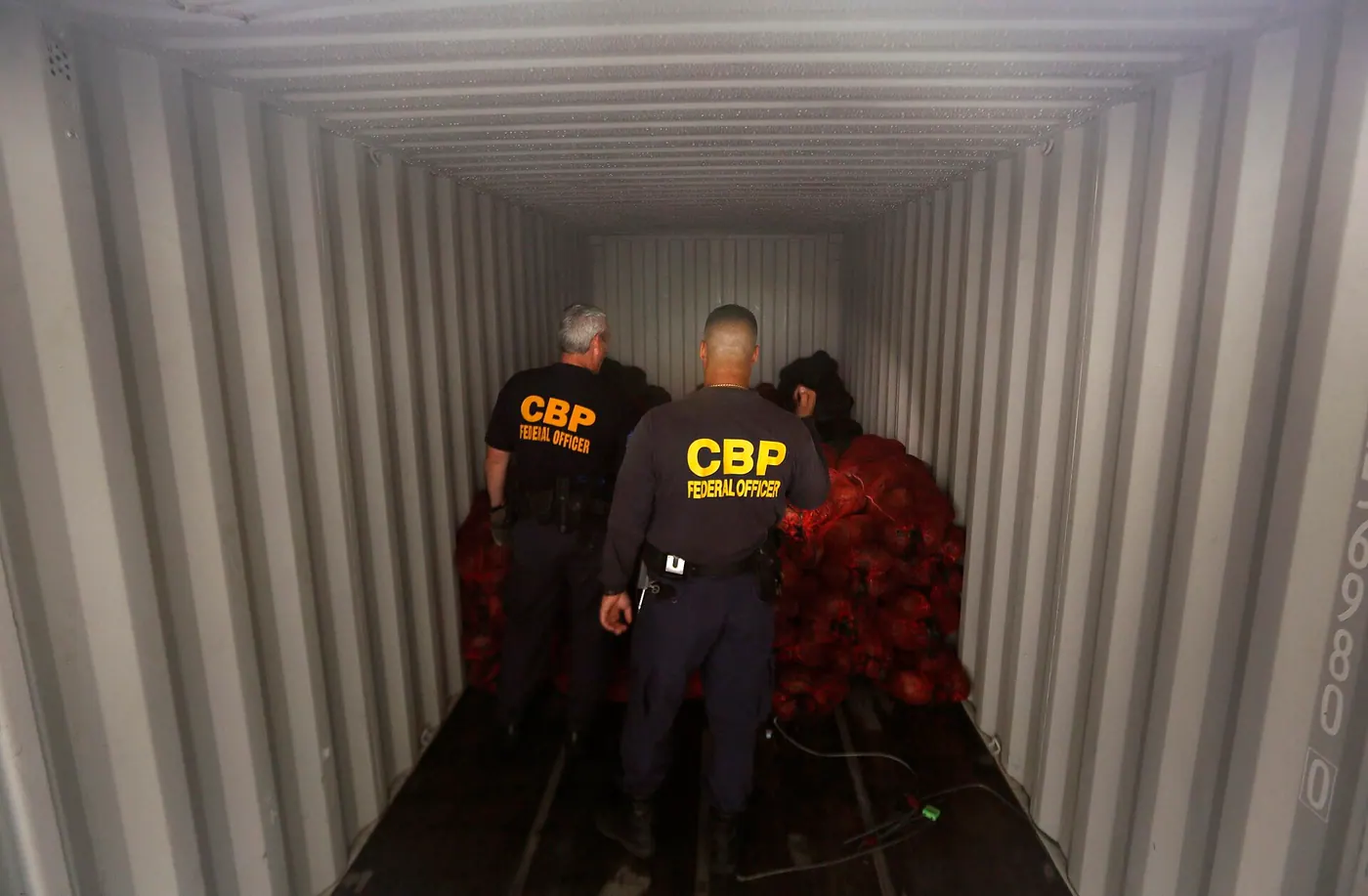 등에 CBP FEDERAL OFFICER 라고 적힌 검은 셔츠 차림의 세관 직원 두 명이 붉은 망 자루가 쌓인 컨테이너 안쪽으로 들어가 있다